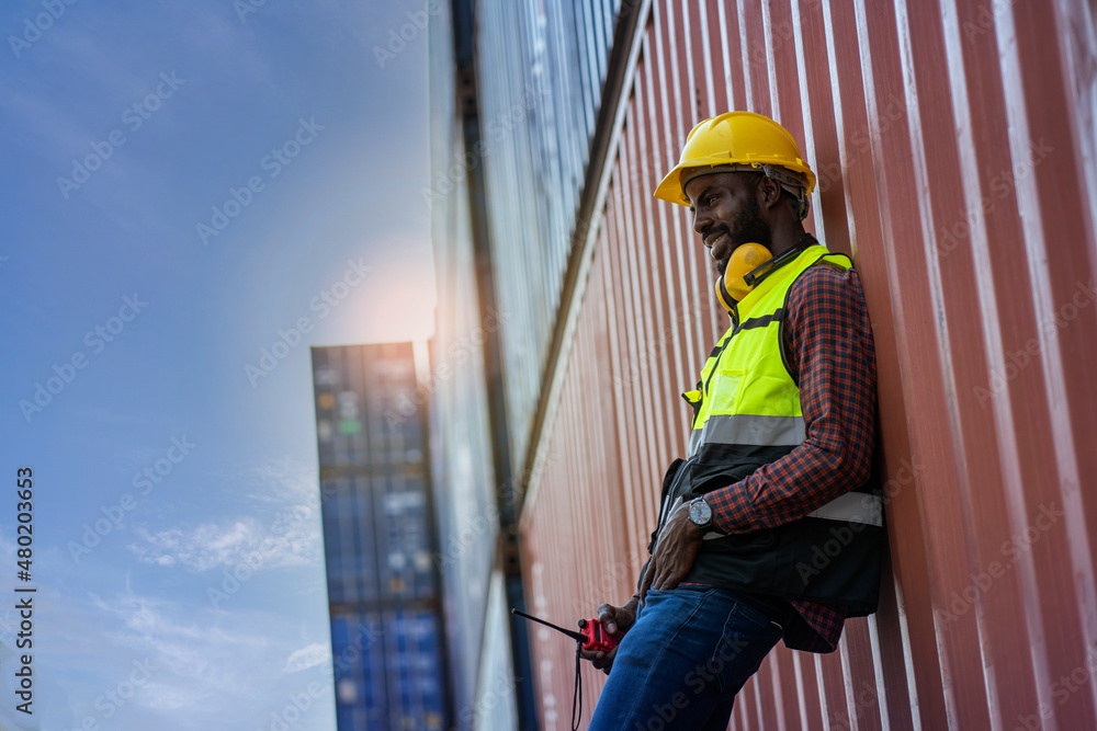 Black worker put uniformed and wearing a safety engineer's hat stands at the container yard at the container yard. Logistics concept . Black worker put uniformed and wearing a safety engineer's hat stands at the container yard at the container yard. Logistics concept .