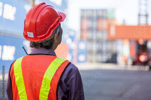Elderly man in a uniform wearing an accident prevention engineer's helmet looks at a container crane at the container yard dock. 