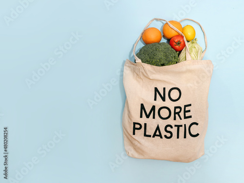 Ecological reusable fabric bag with food. Vegetables and fruits, without plastic. Text on the bag no more plastic. Craft cardboard, cabbage, oranges, tomato, eggs. Banner, free space. Blue background 