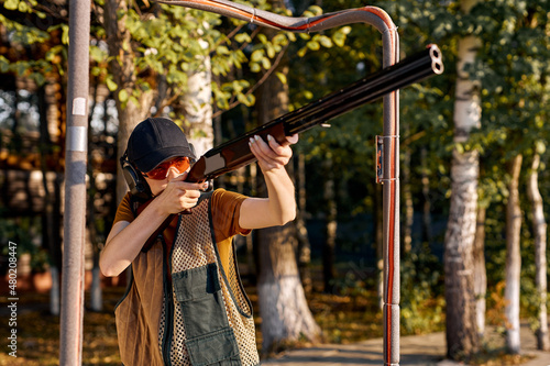 young confident woman in forest standing in the attitude of aiming and looking through the sight of automatic weapon, going to shoot. caucasian lady in protective eyeglasses, cap and headphones.