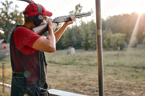 After getting shooting instructions, young caucasian man concentrated at aiming rifle at side, fires at target in outdoor range, wearing goggles, cap and headset equipment outfit. Side view photo