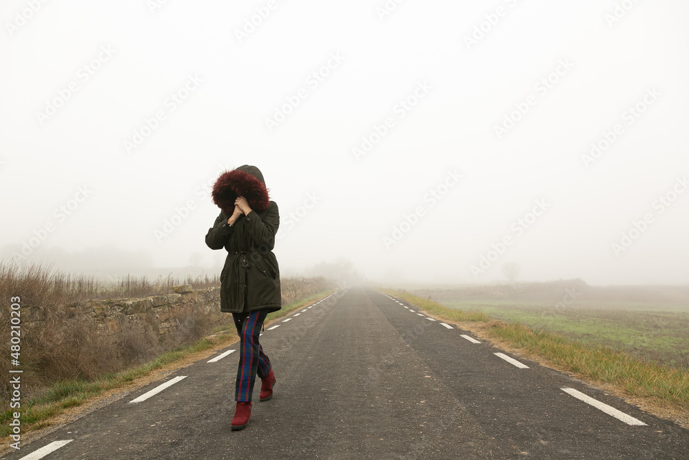 Chica con frío con abrigo caminando con niebla por una carretera secundaria,  huida 