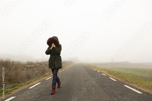 Chica con fr  o con abrigo caminando con niebla por una carretera secundaria   huida 