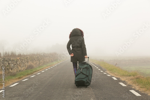 Mujer abrigada en una carretera con niebla huyendo con una bolsa de maleta en solitario, refugiada