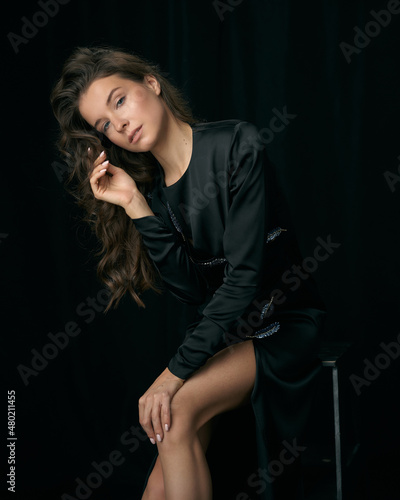 Seductive young woman model with long curly hair and bare legs in elegant cocktail dress sits on bar stool on black background in studio