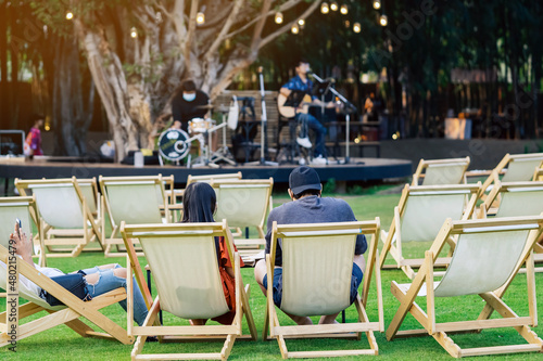 Back view of Asian young couple sit to relax on white deck chairs with tables for dinner in lawn is surrounded by shady green grass with blurred image of musical performance on stage in background.