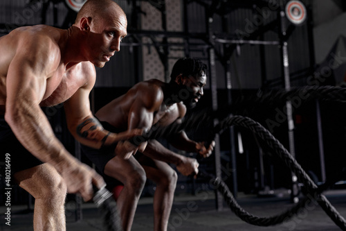 two athletic guys in dark cross fit class, males started training exercises get sweating, holding battle ropes in hands, concentrated and motivated. sportive active healthy lifestyle.