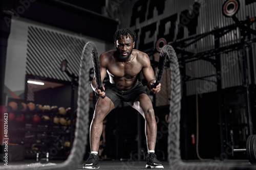 Portrait of strong young african man exercising with battle ropes during workout in modern gym, alone. indoors. active male having intense cross fit training, in dark sports club. fitness concept