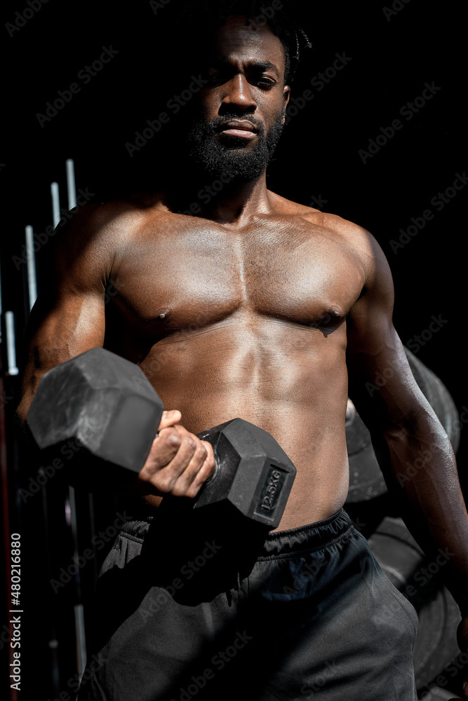 active strong sweaty focused fit muscular man with big muscles holding heavy dumbbell for cross fit training, african man do hard core workout in dark modern gym, real people exercising. portrait