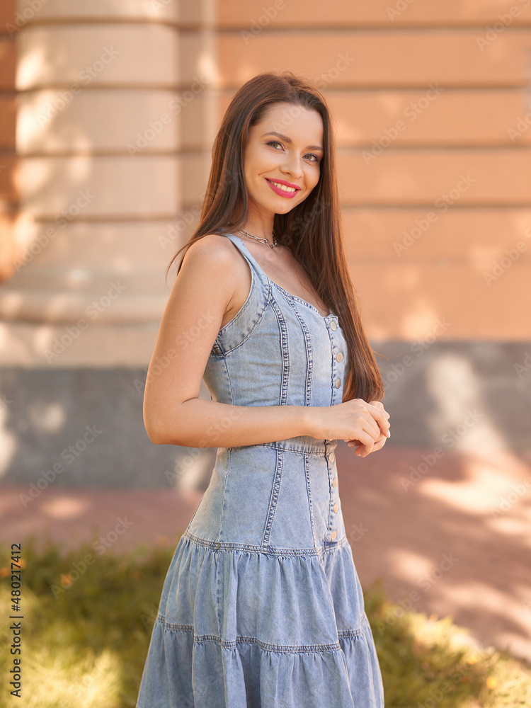 Cheerful long haired brunette lady model in casual denim sundress with red lipstick poses for camera on city street on summer day side view Cheerful long haired brunette lady model in casual denim sundress with red lipstick poses for camera on city street on summer day side view
