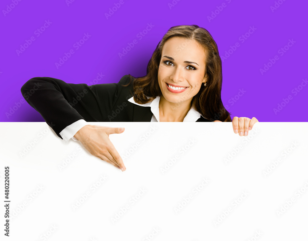 Happy smiling brunette young business woman in blue clothing, standing behind, peeping from blank banner or mock up signboard, showing copy space for text, over purple violet background. Happy smiling brunette young business woman in blue clothing, standing behind, peeping from blank banner or mock up signboard, showing copy space for text, over purple violet background.