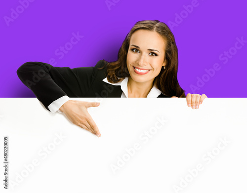 Happy smiling brunette young business woman in blue clothing, standing behind, peeping from blank banner or mock up signboard, showing copy space for text, over purple violet background.