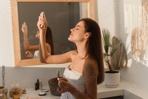 A beautiful young woman, wrapped in a towel, sprays mineral water on her face in front of a mirror. Skin hydration concept.