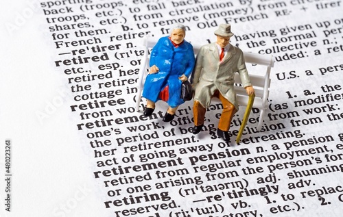 A conceptual image with miniature figures of an old aged elderly couple sitting next to the phrase Retirement Pension on the page of a dictionary