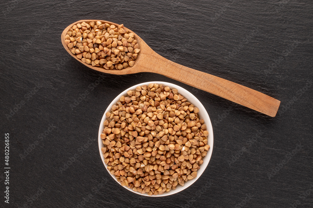 Uncooked organic buckwheat in a white saucer with a wooden spoon on a slate stone, macro, top view. Uncooked organic buckwheat in a white saucer with a wooden spoon on a slate stone, macro, top view.