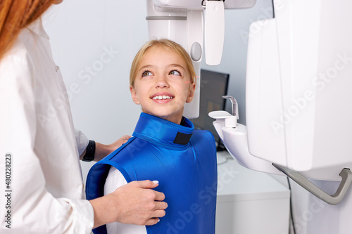 X-ray examination at dental surgery, happy smiling patient in dentist stomatology clinic. Kid girl in blue protective wear sit on chair in front of dental equipment taking x-ray picture. Side view