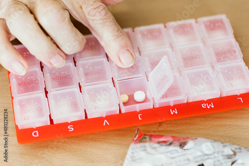 Female hand puts a daily serving of vitamins, medications, tablets, dietary supplements into a red plastic pill organizer or pillbox , close up, toned photo