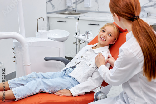 Happy little caucasian child girl in dental surgery waiting on examination couch, side view on professional female redhead dentist calm kid girl down, having nice talk. dental equipment in cabinet