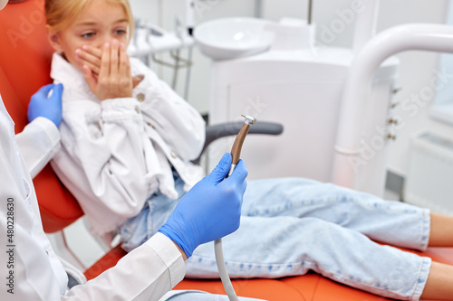 Cropped dentist is going to perform dental filling procedure to frightened child in dental clinic. Doctor using dental tools at work cute kid patient is afraid  having toothache. focus on tool