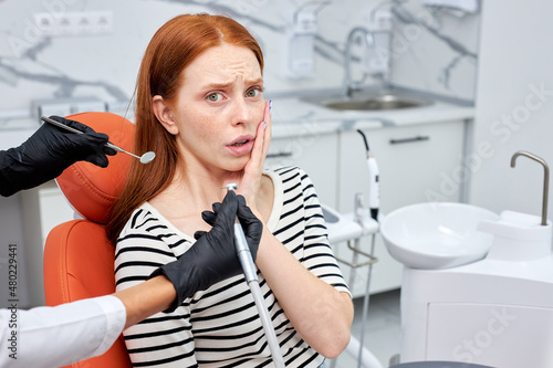 Cropped dentist is performing dental filling procedure to frightened woman in dental clinic. Doctor using dental tools at work  redhead female patient is afraid  having toothache  look at camera