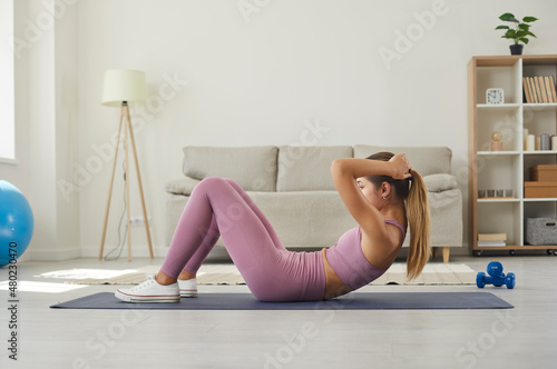 Woman exercising on rubber mat on floor in living room during routine fitness workout at home. Side view of fit young Caucasian girl in sports clothes training her abs by doing sit ups or crunches photo