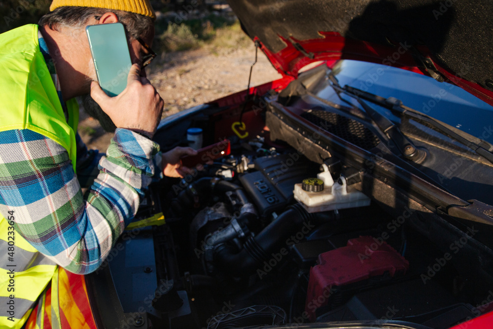 Obraz premium Caucasian man looking under hood of broken down car talking on the phone with mechanic.
