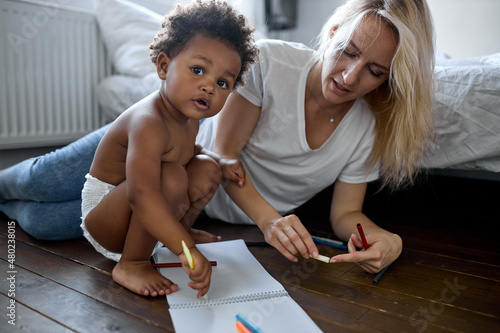 adult mother and small adorable daughter sitting on wooden warm floor at modern home drawing on paper album with colored pencils. Having fun, child development, family leisure activity hobby concept photo