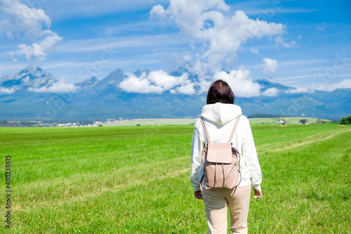 A woman tourist in a white hoodie and a backpack enjoys a beautiful view of the Tatra mountains while standing in a green field. Clouds float over mountain peaks  copy space