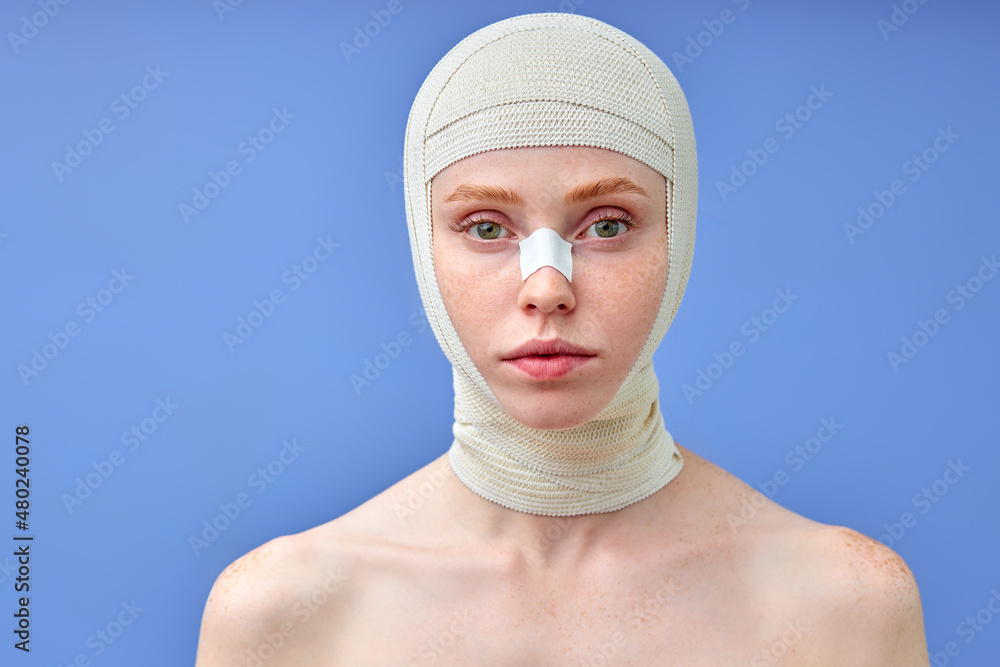 fashionable portrait of young beautiful woman in bandages on an isolated background. caucasian lady posing at camera, after successful but painful nose surgery, isolated on blue. beauty fashionable portrait of young beautiful woman in bandages on an isolated background. caucasian lady posing at camera, after successful but painful nose surgery, isolated on blue. beauty