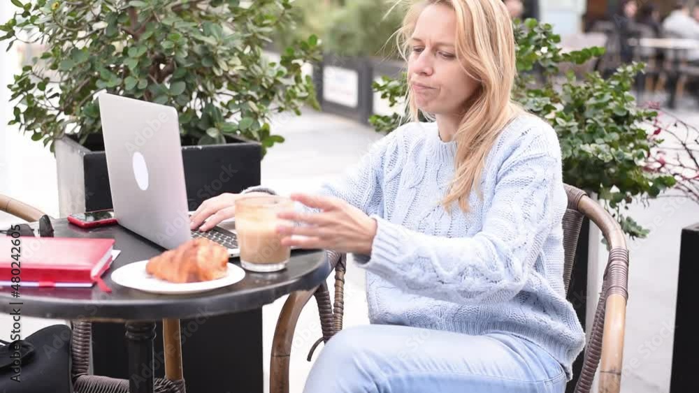 Blonde girl in blue sweater sits on table in cafe with coffee and works on laptop. Thinking face, find solution problem. Work online, freelancer style modern business woman. outdoor Blonde girl in blue sweater sits on table in cafe with coffee and works on laptop. Thinking face, find solution problem. Work online, freelancer style modern business woman. outdoor