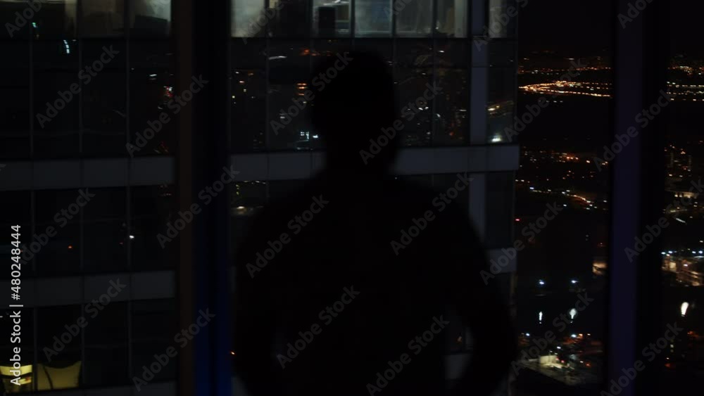 Silhouette of man is standing near the windows in dark office apartment in front of stunning night city urban background on top of skyscraper in modern downtown. Dolly shot camera moving around. Silhouette of man is standing near the windows in dark office apartment in front of stunning night city urban background on top of skyscraper in modern downtown. Dolly shot camera moving around.
