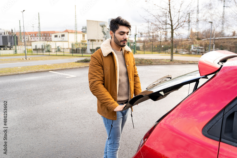 Obraz premium Young latin or arab eastern indian pakistani man taking out or putting stuff in trunk of red rental car on parking slot of urban city getting ready for ride. Travel, car sharing, journey, adventure