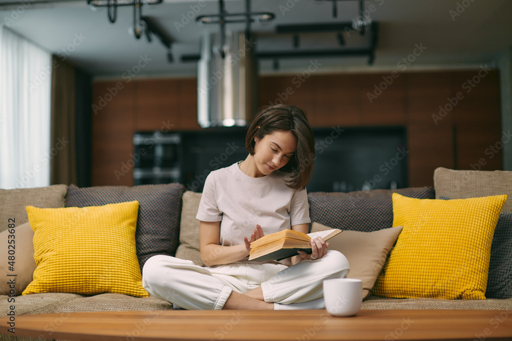 Modern young girl reading bestseller novel in paper book, enjoying weekend, relaxing on sofa at home. Self-education Modern young girl reading bestseller novel in paper book, enjoying weekend, relaxing on sofa at home. Self-education
