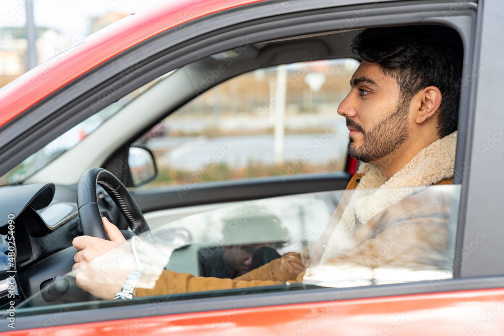 Spanish or arabic ethnicity man in jacket with hands holding driving wheel riding car on autumn, winter or spring day. Travel, exam, lesson, learning, taxi driver. View from outside, opened window