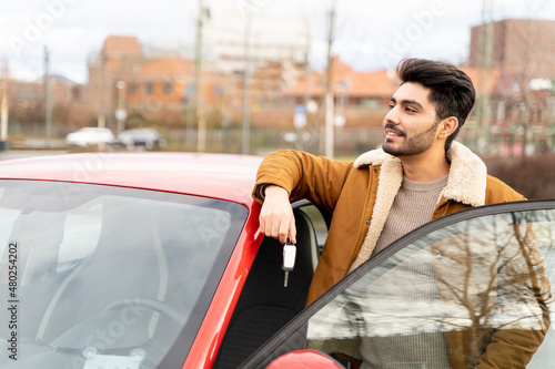 Young latin or arab eastern indian pakistani smiling man in warm clothes holding ignition key standing near red new car with opened door on parking slot of urban city. Travel, rental, car sharing