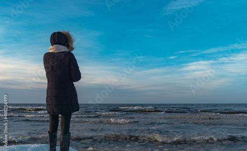 A woman in a black winter jacket and light scarf stands by the sea during the winter. staring into the distance. copy space