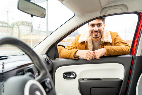 Smiling spanish or arab eastern ethnicity cheerful man in yellow jacket leaned on door looking through window of car as passanger asking for ride or driver. Travel, exam, lesson, learning, taxi driver © pavelgulea