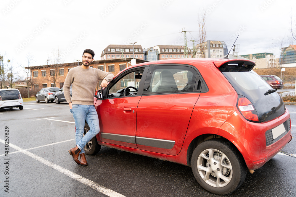 Young latin or arab eastern indian pakistani carefree man in stylish clothes standing near red small car on parking slot of an urban city background on rainy midseason day. Travel, rental, car sharing Young latin or arab eastern indian pakistani carefree man in stylish clothes standing near red small car on parking slot of an urban city background on rainy midseason day. Travel, rental, car sharing