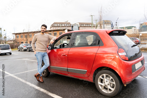 Young latin or arab eastern indian pakistani carefree man in stylish clothes standing near red small car on parking slot of an urban city background on rainy midseason day. Travel, rental, car sharing