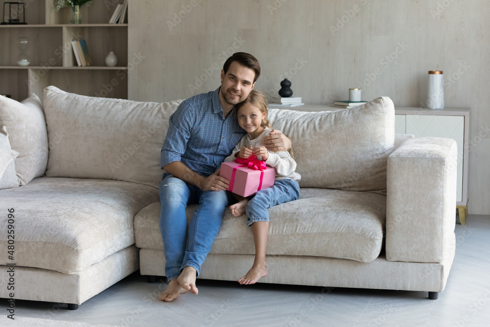 Full length smiling affectionate young father joyful cuddling small cute kid daughter holding wrapped gift box in hands, resting together on sofa at home, cheerful family celebrating happy birthday. Full length smiling affectionate young father joyful cuddling small cute kid daughter holding wrapped gift box in hands, resting together on sofa at home, cheerful family celebrating happy birthday.