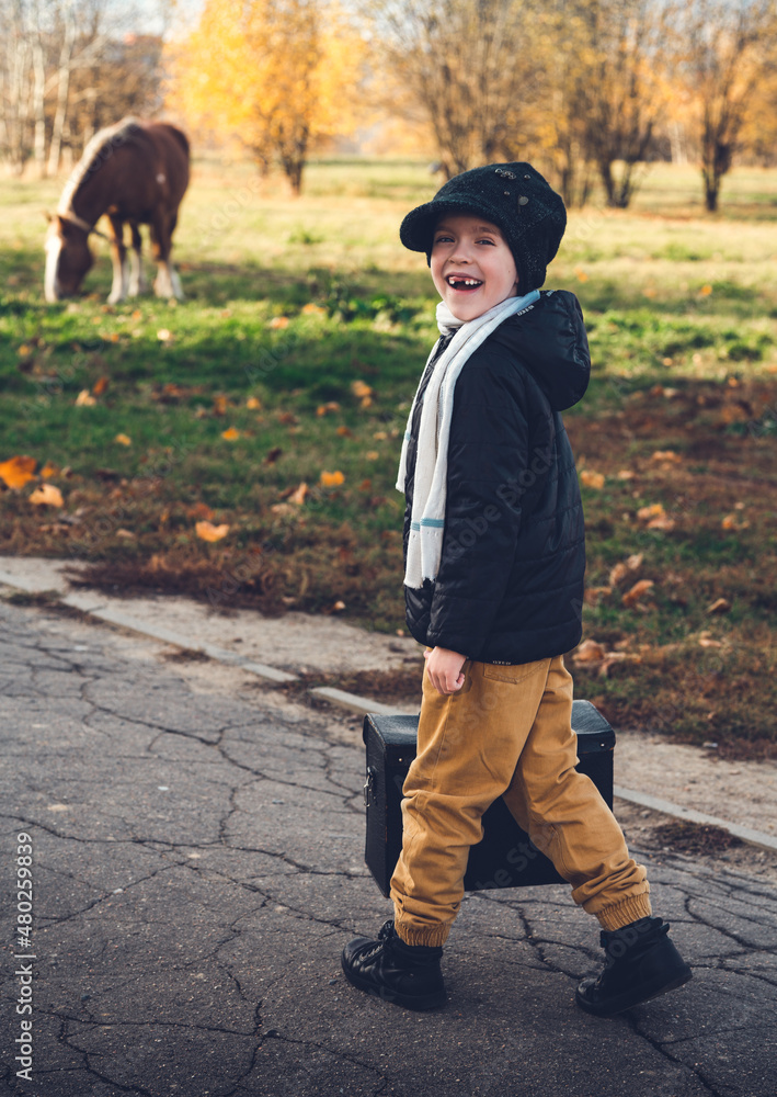 Fototapeta premium A cute little traveler without front teeth smiles, he holds a large leather bag in his hand, steps towards his horse, which is grazing nearby.