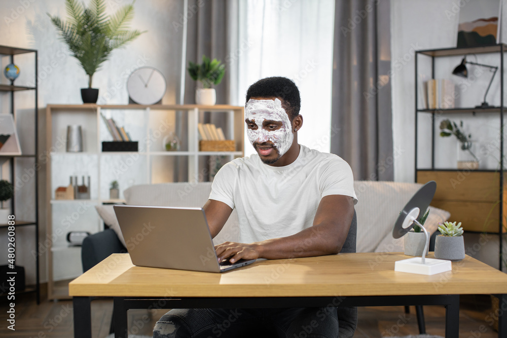 Good-looking smiling young african american man with white facial mask on face typing on laptop, sitting at home and working remotely. Facial mask for skin treatment