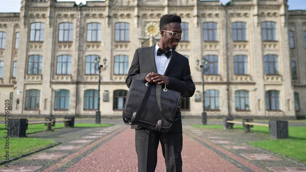 Happy elegant African American man in suit with bowtie rejoicing looking at camera smiling standing outdoors. Portrait of cheerful excited young teacher posing at university campus on sunny morning Happy elegant African American man in suit with bowtie rejoicing looking at camera smiling standing outdoors. Portrait of cheerful excited young teacher posing at university campus on sunny morning