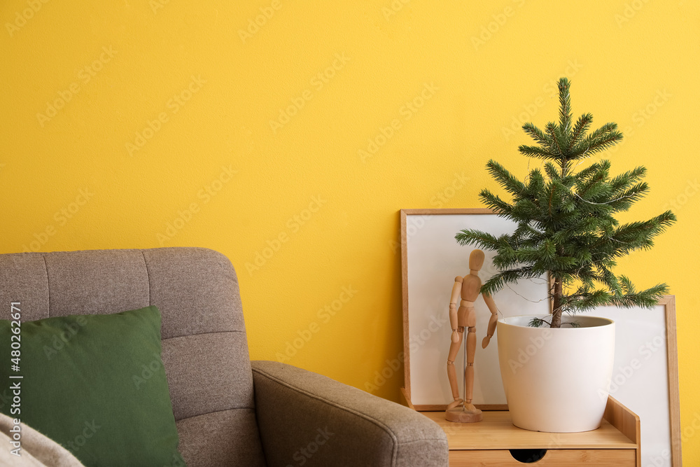 Small Christmas tree in flowerpot on table and armchair near color wall