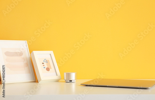 Pictures, portable speaker and modern laptop on table near color wall