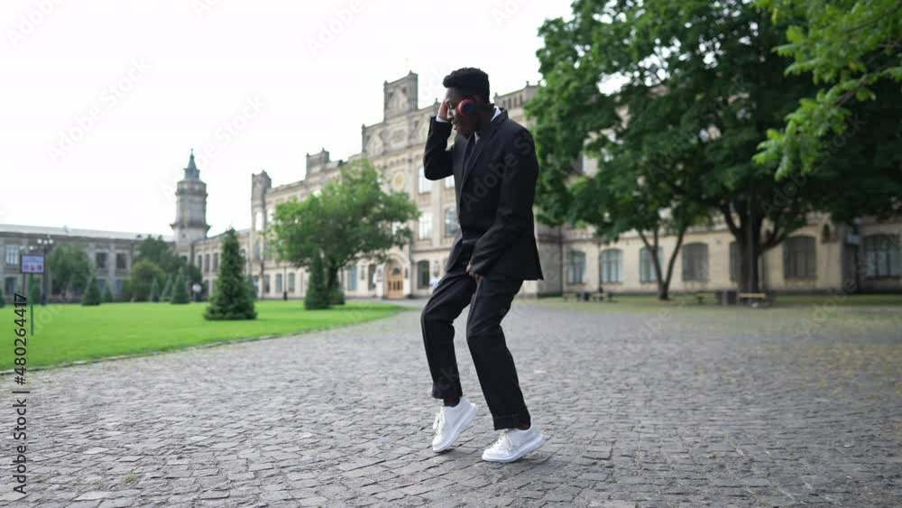 Wide shot relaxed African American joyful student in formal black suit dancing listening to music in headphones on university yard. Happy carefree man having fun enjoying hobby at college campus Wide shot relaxed African American joyful student in formal black suit dancing listening to music in headphones on university yard. Happy carefree man having fun enjoying hobby at college campus