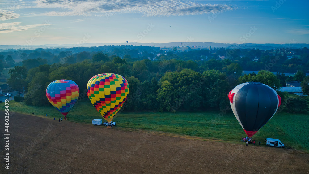 Fototapeta premium Multiple Hot Air Balloons Landing in Farmlands as Amish Look on, and More Still in Flight