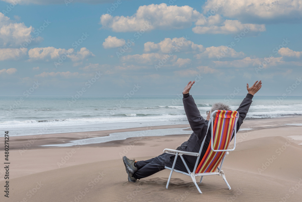 Adult male reclining on a beach lounger in front of the sea with his arms in the air