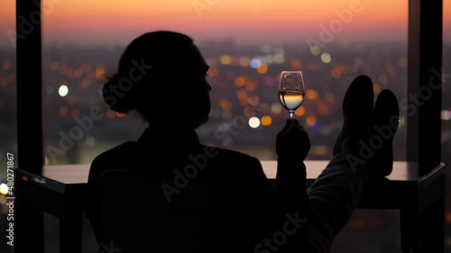 Man sits alone in a dark room by a window, stir and slightly shaking glass of white wine in hand. Lazy evening alone after a busy day at work. Dark city and lights seen blurred on background photo