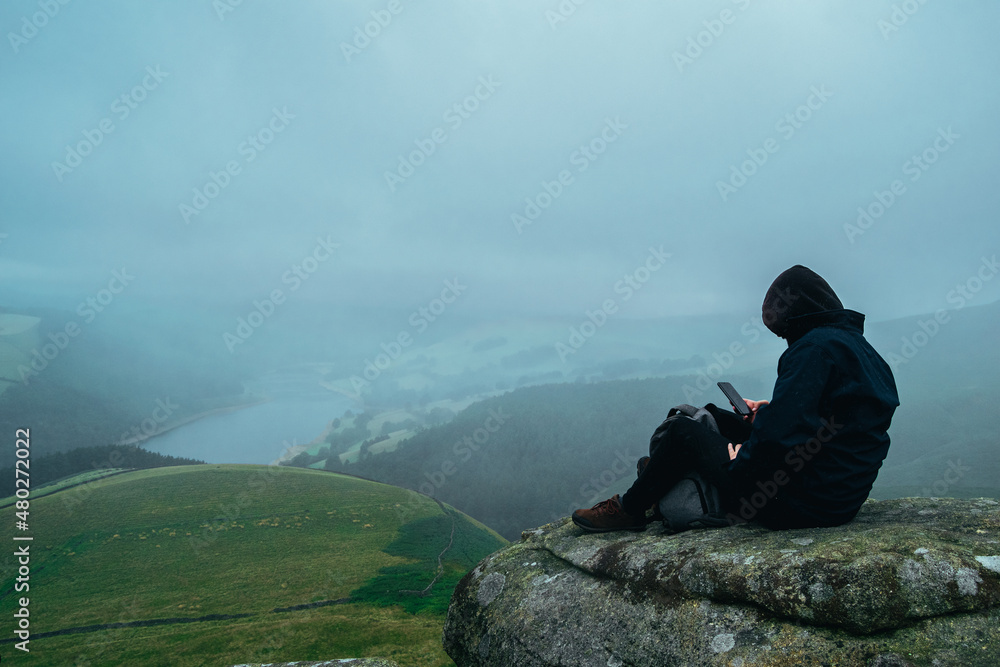 Young male in dark clothes sitting on a rock formation edge looking at phone when surrounded by beautiful landscape. Mobile addiction issue outdoors. Hooded man wearing black browsing on cellphone.